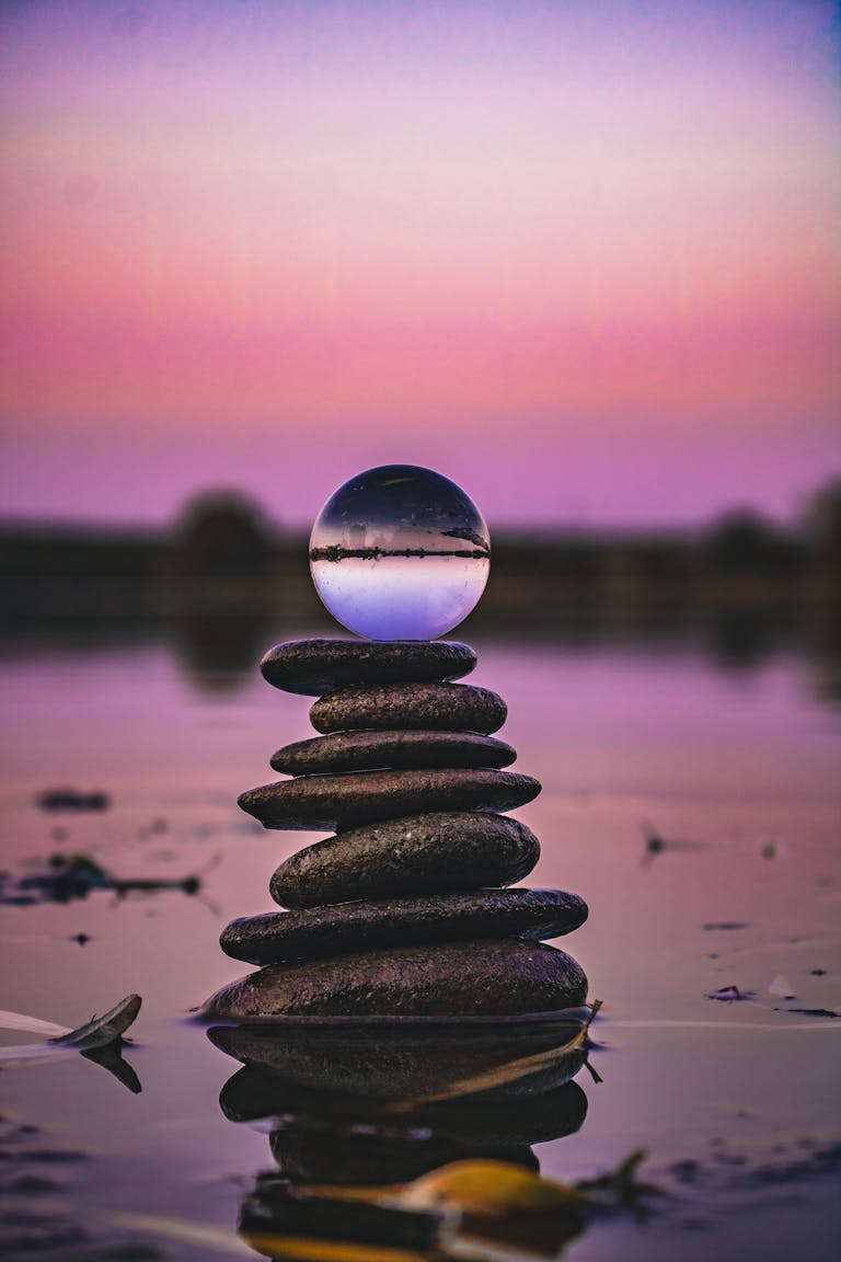Serene image of stacked stones with a glass ball reflection at sunset over water.