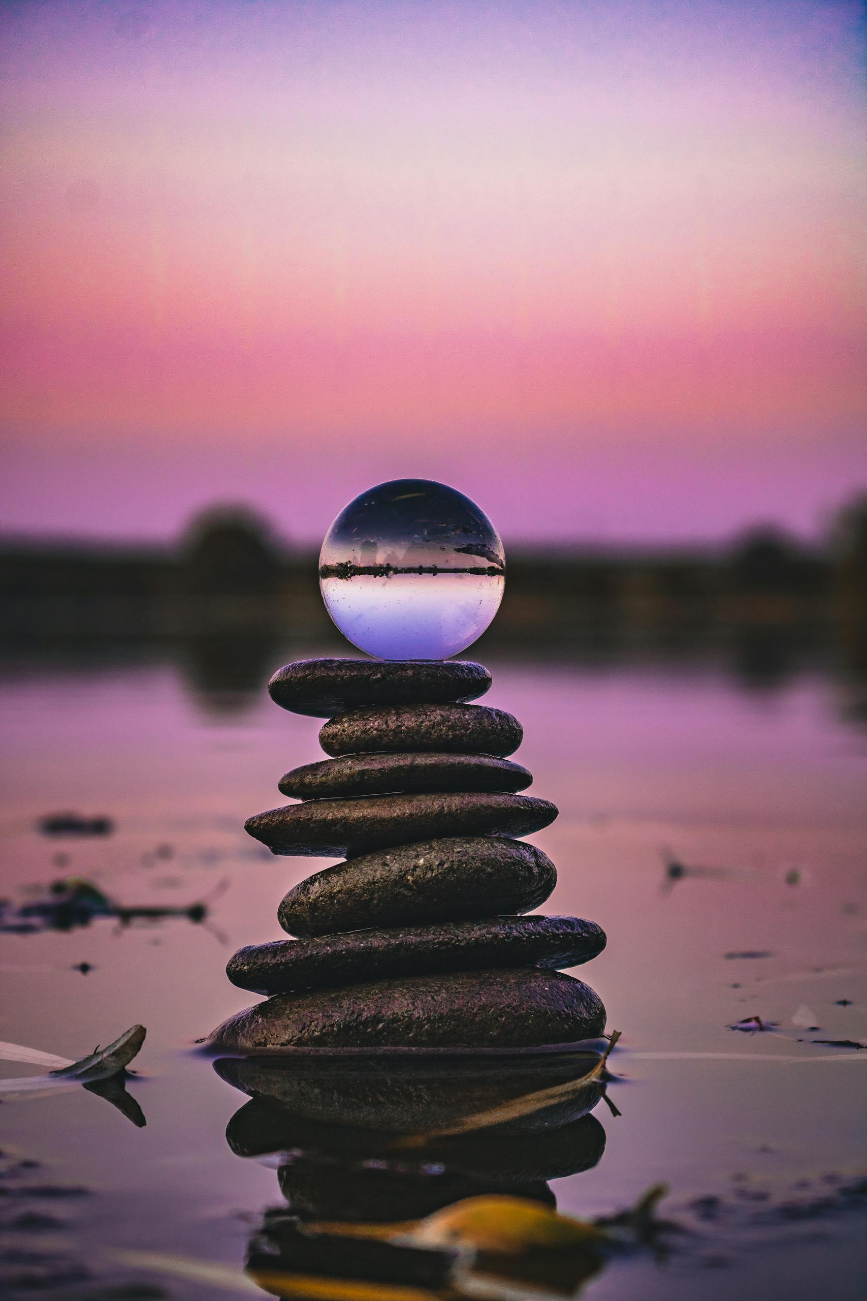 Serene image of stacked stones with a glass ball reflection at sunset over water.